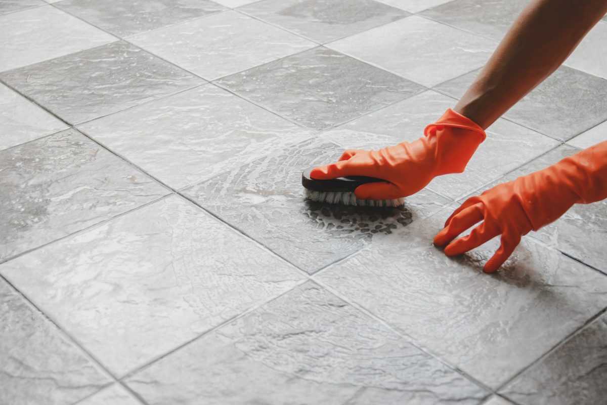 Person in orange rubber gloves scrubbing gray tile floor with brush